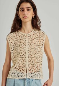 Crochet top in roomkleur met hexagonale patronen, korte mouwen en knoopsluiting, gecombineerd met lichte denim broeken.