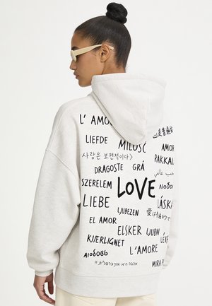 Frau trägt einen weißen Hoodie mit dem Wort "love" in mehreren Sprachen auf dem Rücken und beige Sonnenbrille.