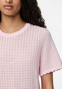 Světle růžový pletený svetr s texturovaným, otevřeným vzorem. Má kulatý výstřih a zvlněné okraje rukávů.
