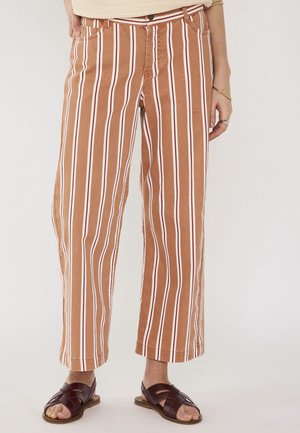 Pantaloni a gamba larga con strisce verticali marroni e bianche indossati con sandali in pelle marrone scuro su sfondo chiaro.