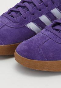 Sneakers de camurça roxa apresentam riscas brancas e solas de borracha gomada, destacando uma forma de bico arredondado com um upper texturizado e costuras reforçadas.