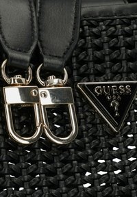Zwarte geweven draagtas met gladde zwarte leren schouderbanden, gouden hardware en een glanzend driehoekig Guess-logo accent.