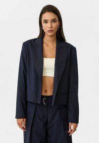 Marineblauw gestreepte cropped blazer met ingezette kraag en voorzakken, gecombineerd met een beige crop top. Het materiaal lijkt glad en gestructureerd.