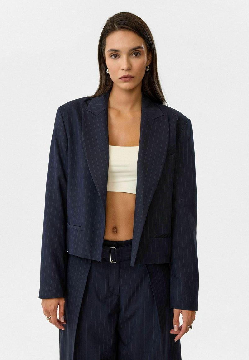 Marineblauw gestreepte cropped blazer met ingezette kraag en voorzakken, gecombineerd met een beige crop top. Het materiaal lijkt glad en gestructureerd.
