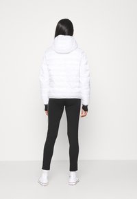 Giacca a vento bianca con cappuccio, caratterizzata da un design trapuntato. Abbinata a leggings neri e sneakers bianche. Nessun marchio o logo visibile.