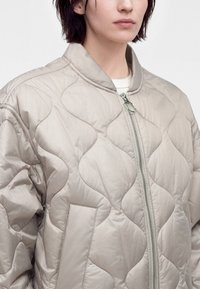 Personne portant une veste matelassée beige clair avec des surpiqûres en forme de vague, une fermeture éclair à l’avant, un col et des poches latérales visibles, sur un fond uni.