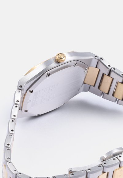 RVS horloge met een gouden accent, octagonale behuizing, metalen band met zilveren en gouden schakels, zichtbare kroon, gedetailleerde gravure op de achterkant.