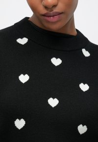 Maglione nero con scollo tondo, caratterizzato da motivi a cuori bianchi distribuiti uniformemente su tutto il tessuto. Tessuto morbido e design a maglia.