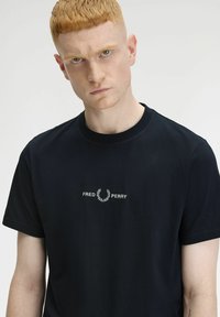 T-shirt nera in cotone con colletto girocollo e maniche corte, decorata con il logo "Fred Perry" ricamato in bianco e dettagli di alloro sul petto.