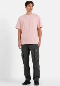 Lichtroze katoenen T-shirt met korte mouwen, voorzien van subtiele witte tekst. Gecombineerd met donkergrijze cargobroek met meerdere zakken.