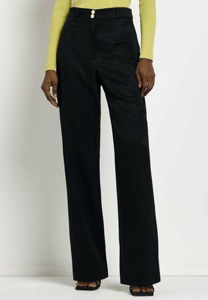 Modelka má na sobě černé kalhoty s vysokým pasem a širokými nohavicemi se zapínáním na dvojité knoflíky, doplněné žlutým dlouhým rukávem a černými sandály s otevřenou špičkou.