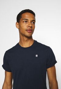 Dunkelblaues Baumwoll-T-Shirt mit kurzen Ärmeln und umgeschlagenen Bündchen. Verfügt über ein kleines weißes Logo auf der Brust. Glatte Textur, klassischer Rundhalsausschnitt.