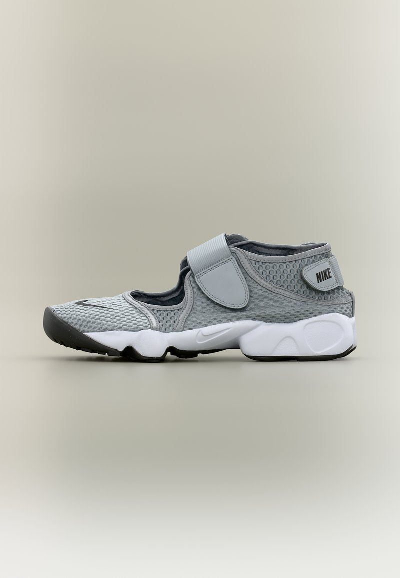 Αθλητικό παπούτσι Nike σε ανοιχτό γκρι χρώμα με επάνω μέρος από δίχτυ, ρυθμιζόμενους ιμάντες και λευκή μαλακή σόλα, σε ουδέτερο φόντο.