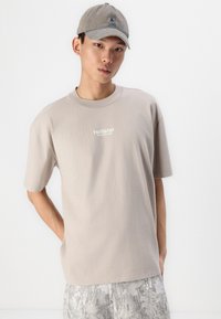 Graues Baumwoll-T-Shirt mit kurzen Ärmeln, ausgestattet mit einem kleinen weißen "Hollister"-Logo auf der Brust, kombiniert mit hellen, gemusterten Shorts und einer passenden Mütze.