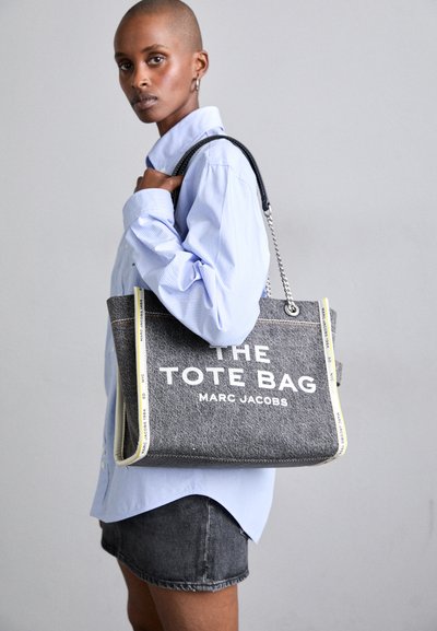 Sac fourre-tout en toile grise avec texte blanc, quincaillerie argentée et accents jaunes. Dispose de poignées robustes et d'un design spacieux pour transporter des objets.