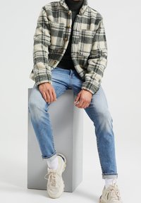 Fleecetakki kermaan ja vihreään ruutukuvioon, jossa puiset napit ja kaulus, yhdistetty vaaleansinisiin farkkuihin ja paksupohjaisiin beigen sävyisiin lenkkareihin.