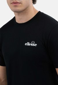 Mężczyzna w czarnej koszulce z krótkim rękawem z białym logo "ellesse" na lewej piersi, na jednolitym jasnym tle.