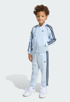 Light blue tracksuit made of smooth fabric featuring dark blue side stripes. Includes zip-up jacket and matching pants, with logo accents.