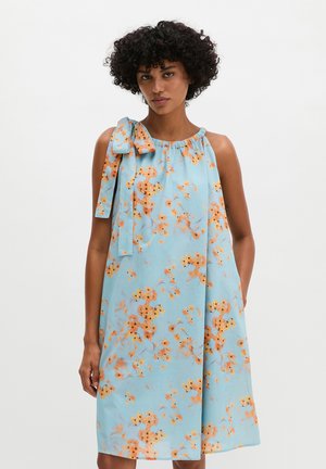 Frau mit lockigem Haar, die ein ärmelloses hellblaues Kleid mit orangefarbenem Blumenmuster und einer Schleife an der Schulter trägt, steht vor weißem Hintergrund.