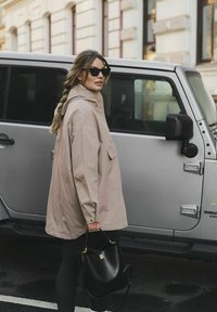 Žena v béžovém kabátu a slunečních brýlích stojí vedle stříbrného SUV a drží černou kabelku na městské ulici.