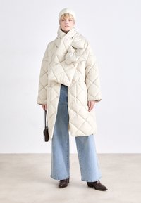 Casaco de puffer acolchoado em creme com silhueta oversized. Combinado com jeans de perna larga em azul claro e botas de tornozelo castanhas escuras. Gorro de malha e cachecol.
