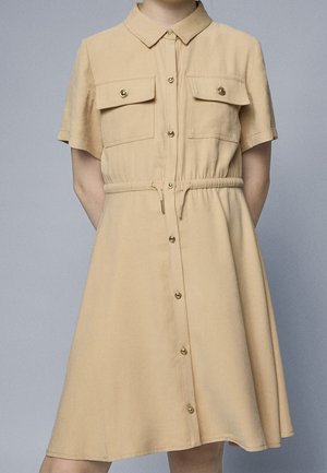 Beige jurk met korte mouwen, een knoopsluiting aan de voorkant, twee borstzakken, een taillekoord en gouden sierdetails. Gladde stoftextuur.