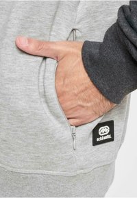 Sweatshirt gris avec une poche zippée, arborant un patch avec logo noir. Le tissu a une texture lisse et la manche est d'un gris plus foncé.
