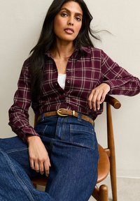 Femme aux longs cheveux noirs assise sur une chaise en bois, portant une chemise à carreaux bordeaux à boutons, un jean taille haute bleu, et une ceinture marron avec une boucle dorée.