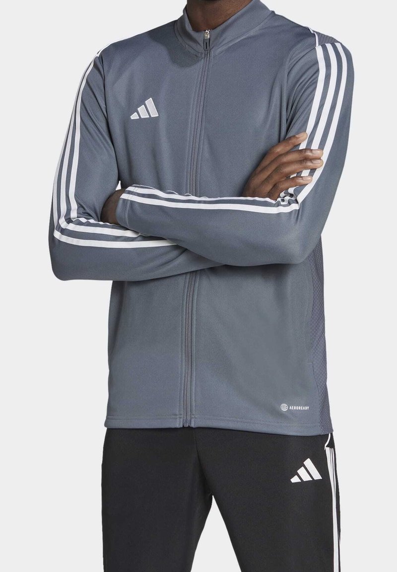 Veste de sport grise en matériau qui évacue l'humidité, avec un col haut, une fermeture éclair à l'avant et des accents rayés blancs sur les manches.