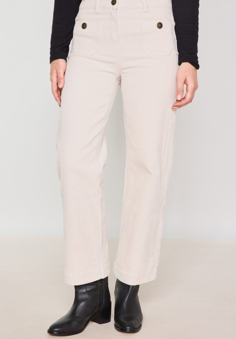 Beige fluwelen wijde broek met twee vo pockets, hoge taille en twee knopen, gecombineerd met zwarte enkellaarzen, staand op een neutrale achtergrond.
