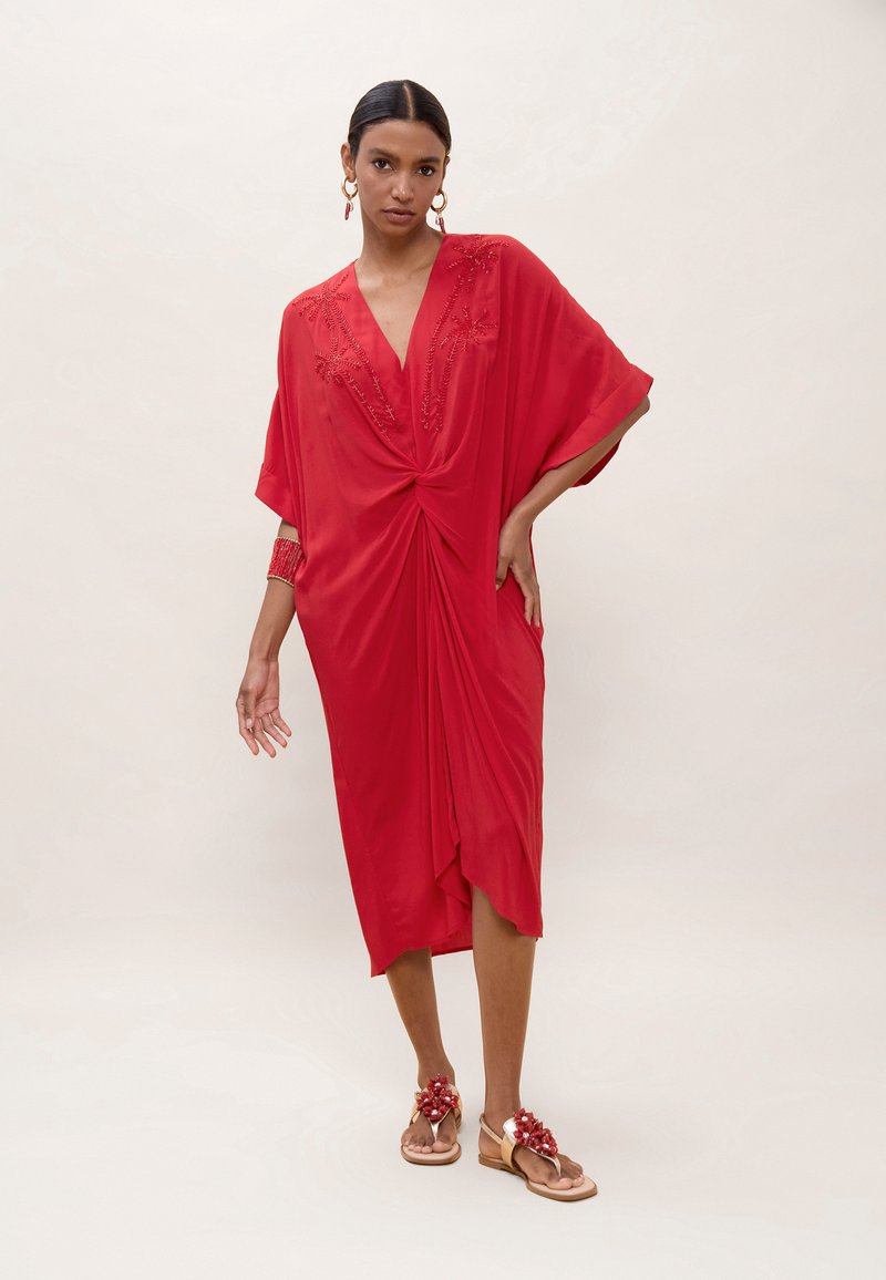 Femme debout portant une robe rouge mi-longue ornée de perles, avec un détail torsadé à l'avant, des boucles d'oreilles créoles dorées, un bracelet rouge et des sandales à fleurs.