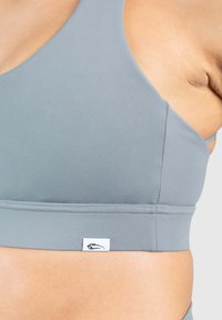 Szary stanik sportowy wykonany z gładkiego, rozciągliwego materiału. Posiada szerokie ramiączka, bezszwowy design oraz małą metkę z logo przy dolnej krawędzi.