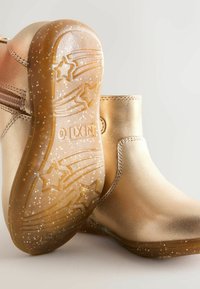 Botas de tobillo metálicas doradas con un acabado suave, que cuentan con una cremallera lateral y una suela de goma texturizada y brillante adornada con patrones de estrellas.