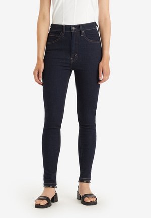 Vrouw draagt donkerblauwe high waist skinny jeans met contrasterende stiksels en zwarte sandalen met blokhak, staand tegen een witte achtergrond.