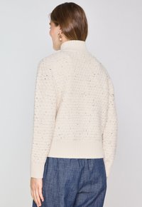 Pull en crème tricoté avec une surface texturée, col montant et poignets et ourlet côtelés. Associé à un pantalon bleu foncé au tombé décontracté.