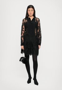 Svart gjennomsiktig bluse med lange ermer, kombinert med et lagdelt svart blondeskjørt. Modellen har på seg svarte strømpebukser og holder en liten svart håndveske.