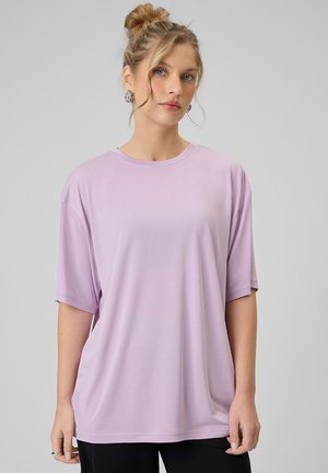 Młoda kobieta o blond włosach związanych w kok, nosząca srebrne kolczyki koła oraz luźną, jasnofioletową koszulę z krótkim rękawem na jednolitym szarym tle.