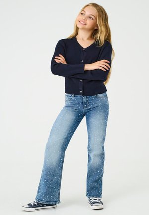Marineblå ribstrikket cardigan med knapper, kombineret med lyseblå jeans med et spraglet mønster. Afsluttet med marineblå sneakers.