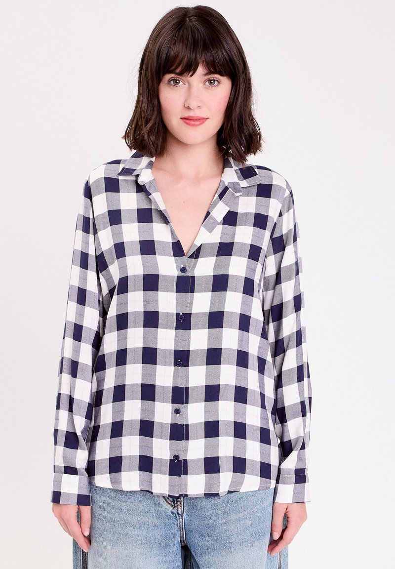 Chemise à manches longues et col en V à carreaux, avec un motif à damier bleu marine et blanc, devant à boutons et en tissu doux. Associée avec un jean bleu clair.