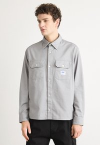 Gray button-up shirt made of cotton, featuring a collar, two front pockets, and a small logo tag. Long sleeves with buttoned cuffs.