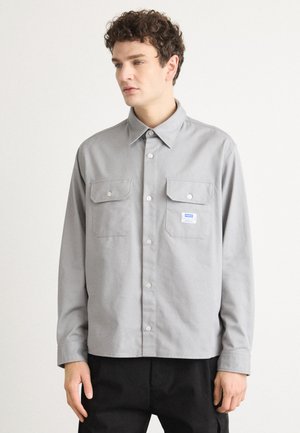 Gray button-up shirt made of cotton, featuring a collar, two front pockets, and a small logo tag. Long sleeves with buttoned cuffs.