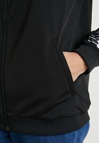 Svart zip-jacka med en slät yta, med sidofickor och randig detalj på ärmen. Stretchig nederkant och ärmslut.