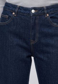 Σκούρα μπλε τζιν denim με πορτοκαλί ραφές φορεμένα με λευκό ριμπ πλεκτό μπλουζάκι που είναι μαζεμένο στη μέση.