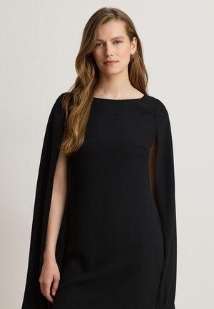 Une femme porte une robe noire à manches cape sur un fond clair et uni.