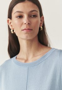 Maglione azzurro chiaro con scollatura rotonda e rifiniture a coste. Il tessuto sembra morbido, con una sottile trama e un design semplice. Orecchini ad anello dorati.