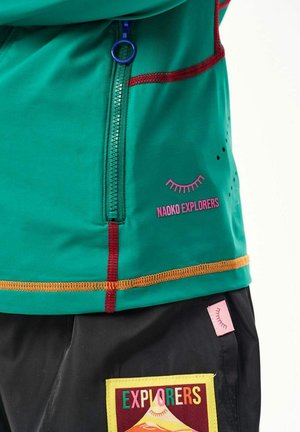 Turkusowa kurtka z czerwonym i pomarańczowym przeszyciem, niebieskim zamkiem błyskawicznym, napisem „NAOKO EXPLORERS” oraz czarne spodnie z kolorową naszywką „EXPLORERS” i różową metką.