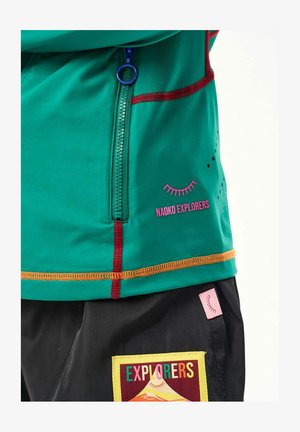 Turkusowa kurtka z czerwonym i pomarańczowym przeszyciem, niebieskim zamkiem błyskawicznym, napisem „NAOKO EXPLORERS” oraz czarne spodnie z kolorową naszywką „EXPLORERS” i różową metką.
