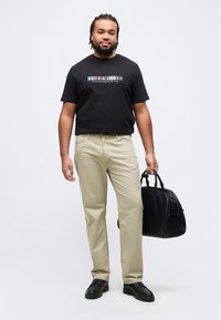 Homme avec des cheveux tressés portant un t-shirt noir à motifs, un pantalon beige, des chaussures noires, tenant un sac en cuir noir sur un fond blanc.