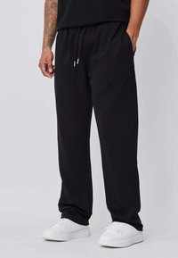 Pantalon de jogging noir en tissu doux, avec une ceinture élastique à cordon, des poches latérales et une coupe décontractée. Porté avec des baskets blanches.