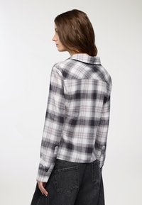 Camicia a quadri in grigio, bianco e nero; maniche lunghe; colletto button-down; texture morbida; indossata con una gonna nera.